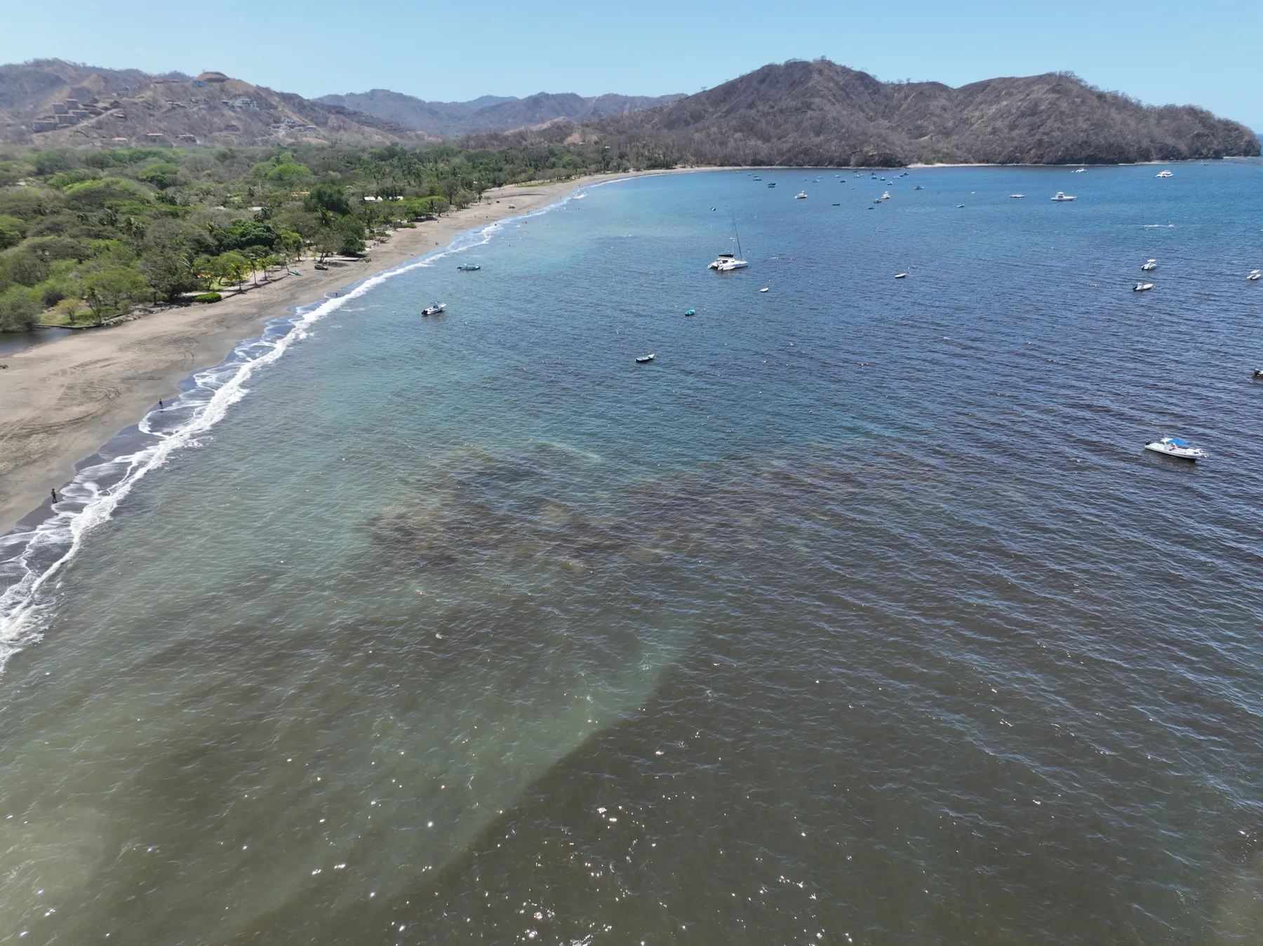 Aerial view of a curving Guanacaste beach and a calm bay with small boats at anchor, dry forested hills behind
